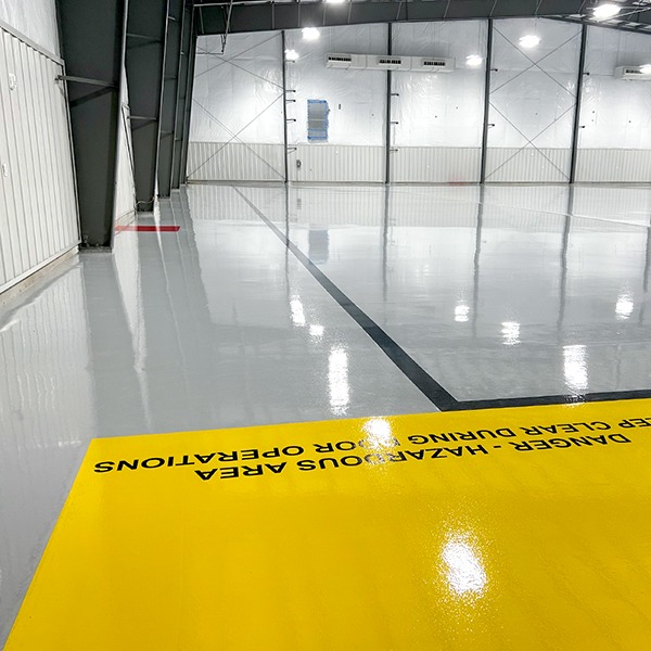 Large industrial warehouse with glossy, polished concrete floors, yellow safety zone marked "DANGER HAZARDOUS AREA," and white paneled walls with overhead lighting.