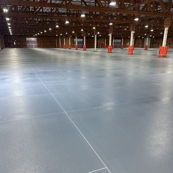 Large, empty warehouse with polished concrete floors, exposed beams, and rows of columns wrapped with red padding under bright overhead lighting.