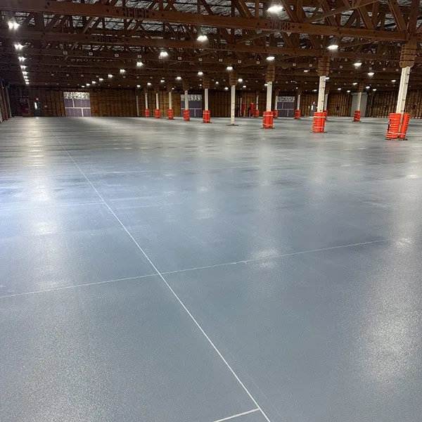 Large, empty warehouse with polished concrete floors, exposed beams, and rows of columns wrapped with red padding under bright overhead lighting.
