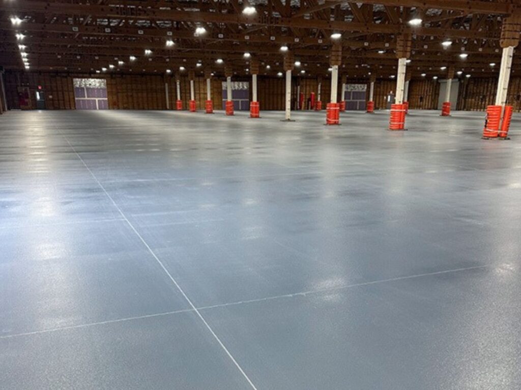 Large empty warehouse with polished concrete floors, white support columns, exposed wooden ceiling beams, and orange barriers spaced throughout.