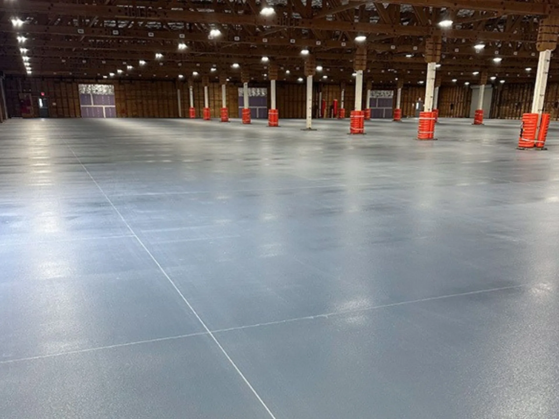 Large empty warehouse with polished concrete floors, white support columns, exposed wooden ceiling beams, and orange barriers spaced throughout.