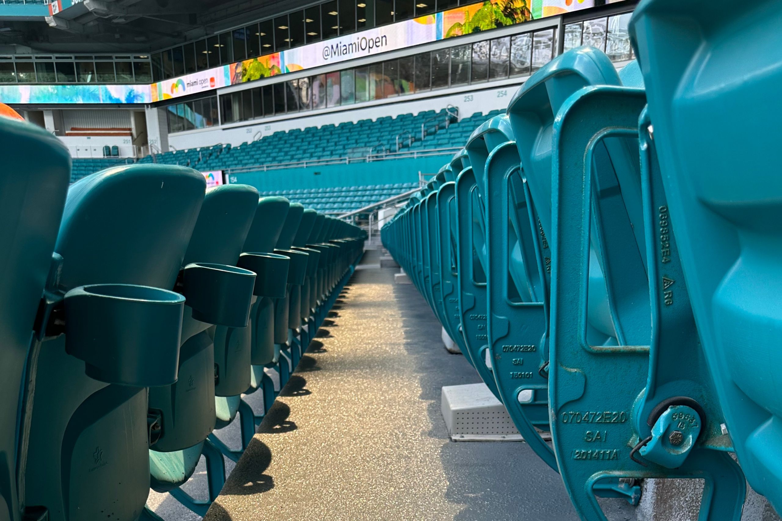 Rows of empty teal stadium seats with cup holders, photographed from aisle level; sturdy commercial and industrial flooring lines the walkways as a digital sign above displays "@MiamiOpen.