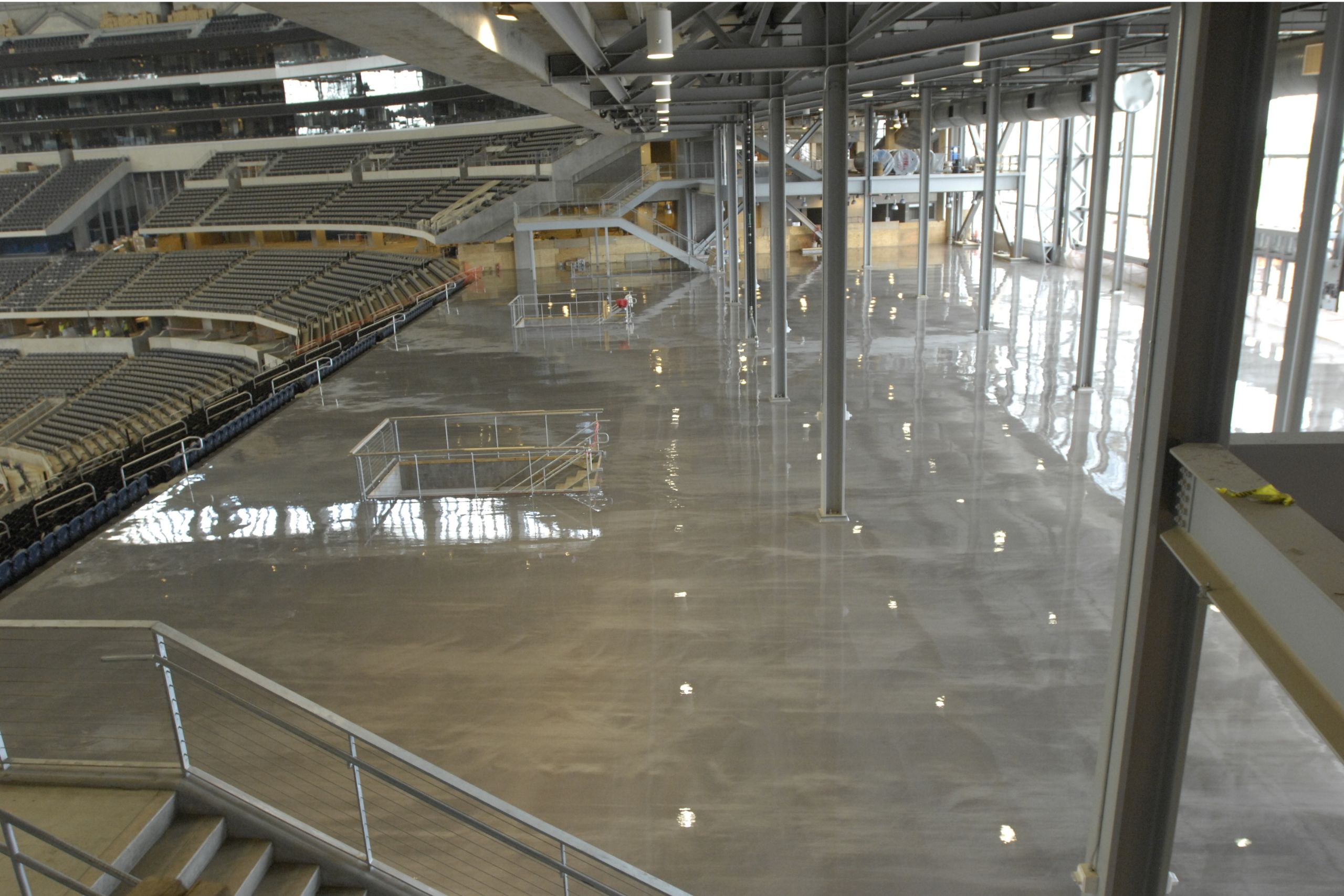 Large indoor stadium area featuring commercial and industrial flooring with polished concrete, metal railings, empty seating sections, and visible overhead structural beams.
