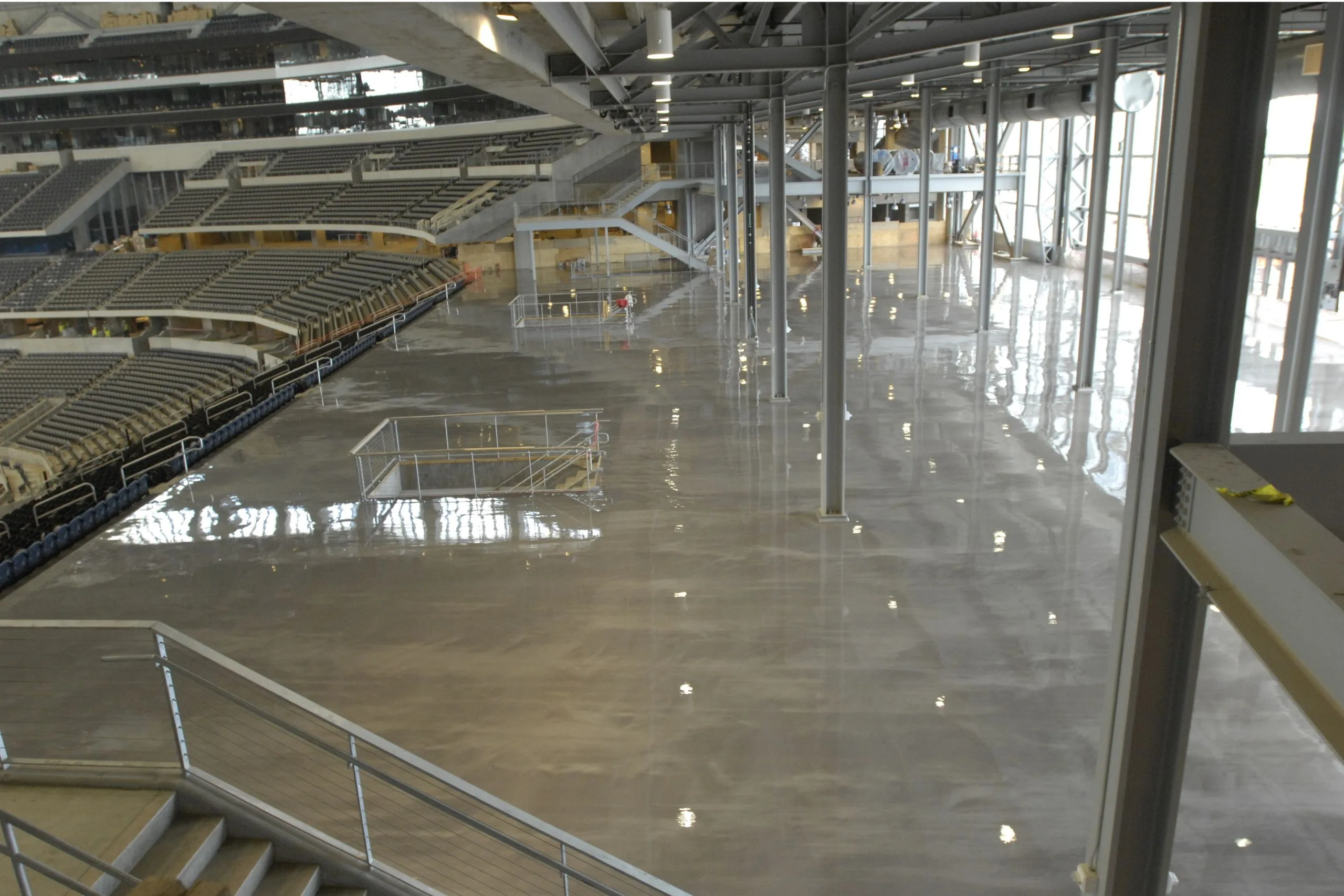 Large indoor stadium area featuring commercial and industrial flooring with polished concrete, metal railings, empty seating sections, and visible overhead structural beams.