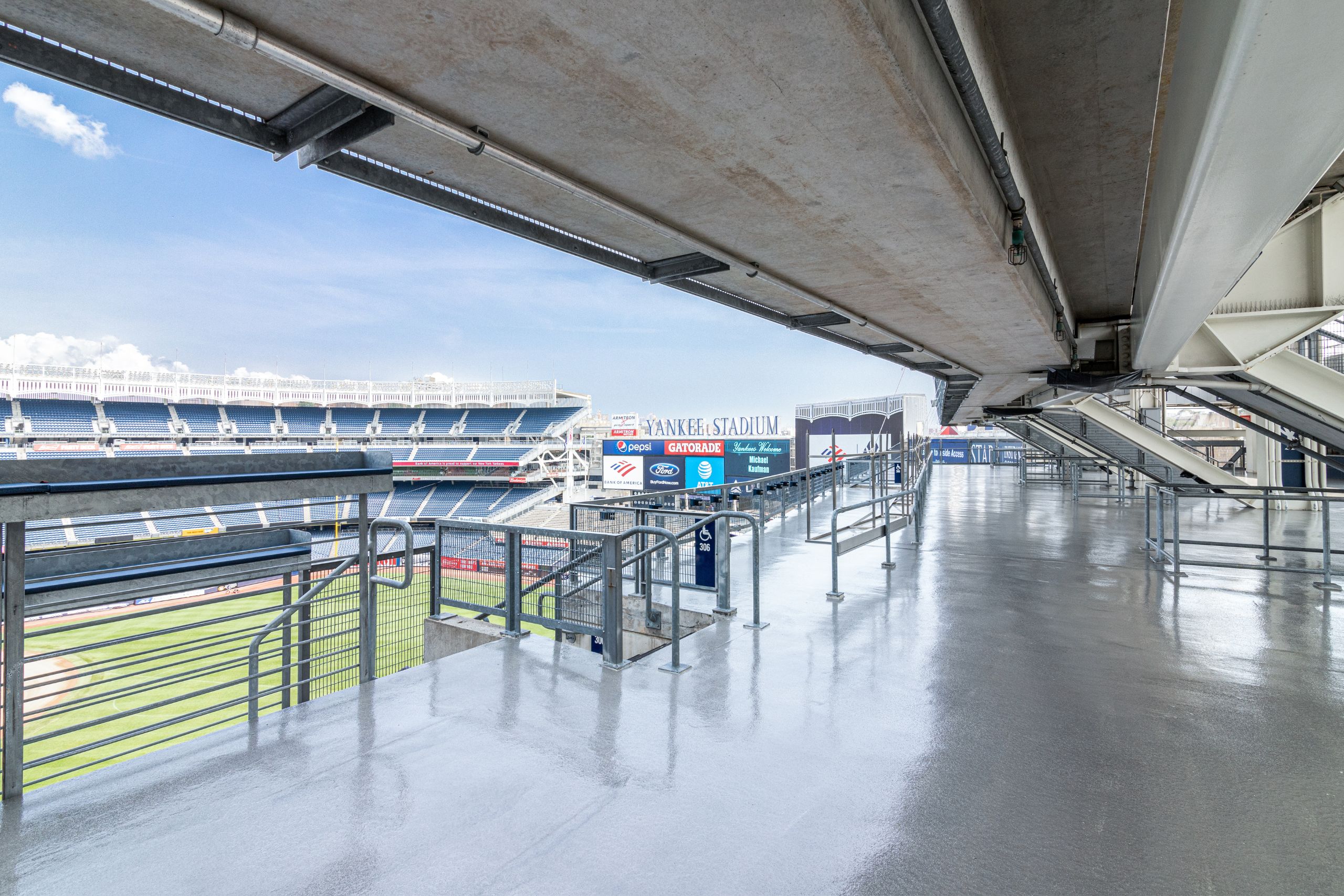 View of an empty concourse inside Yankee Stadium, featuring commercial and industrial flooring, railings, signage, and a glimpse of the baseball field under a clear sky.