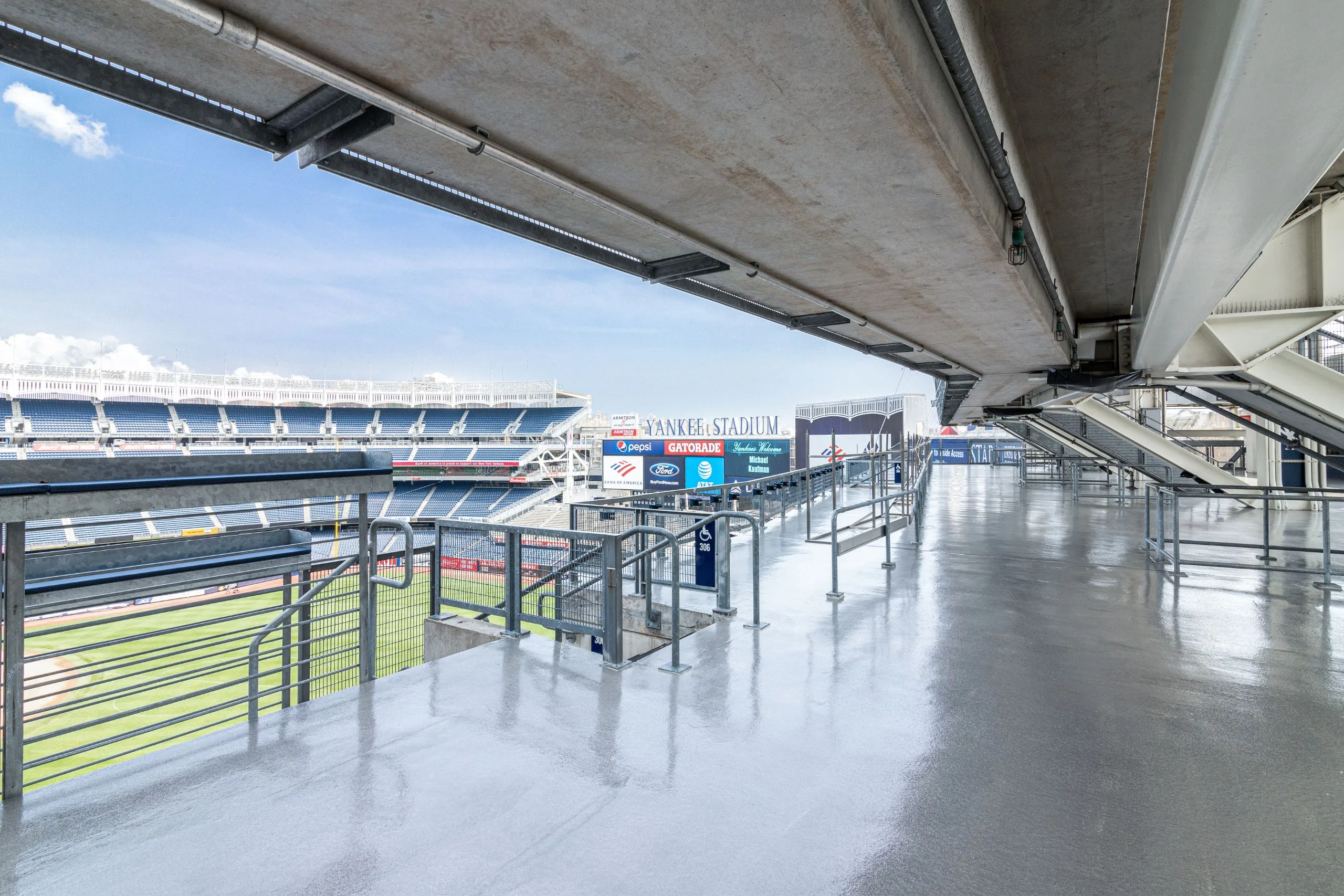 View of an empty concourse inside Yankee Stadium, featuring commercial and industrial flooring, railings, signage, and a glimpse of the baseball field under a clear sky.