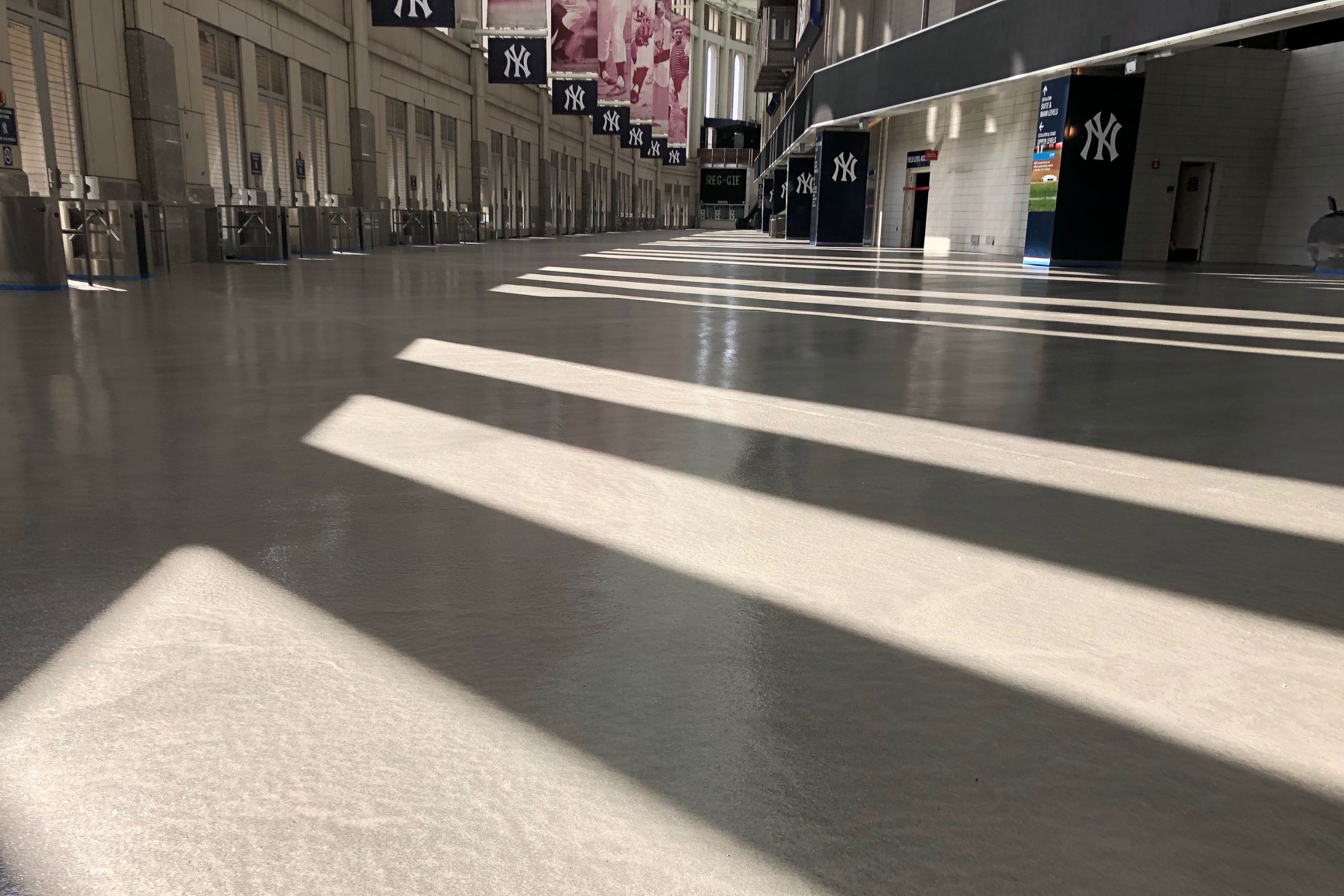 Sunlight casts diagonal shadows on the commercial and industrial flooring of an empty concourse lined with New York Yankees banners and closed concession stands.