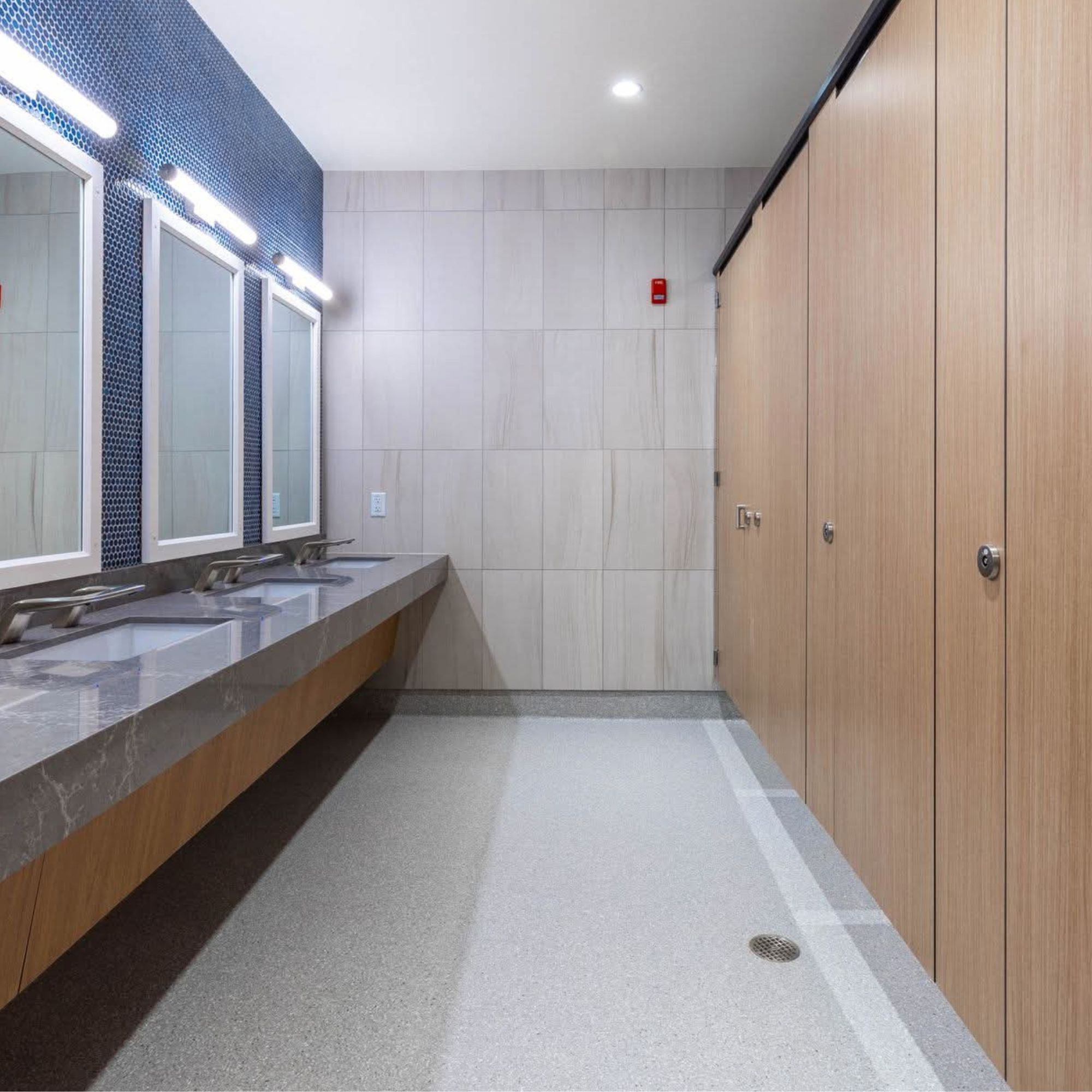 Modern public restroom with three sinks and mirrors on the left, beige tiled walls, and wooden stall doors on the right. The floor is light-colored with a drain in the center.