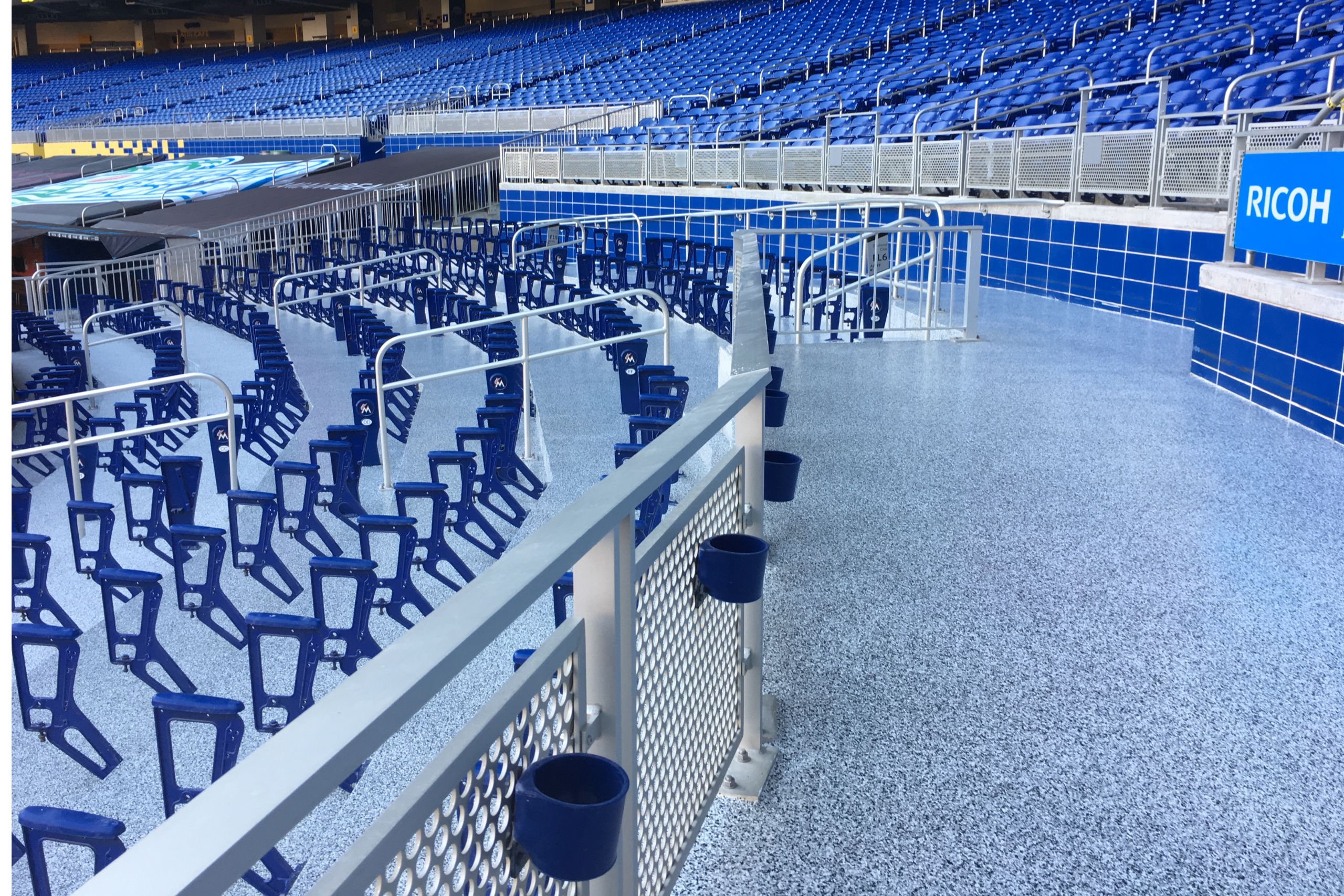 Rows of empty blue stadium seats are seen behind metal railings inside a large, modern arena with blue accents, signage, and durable commercial and industrial flooring.