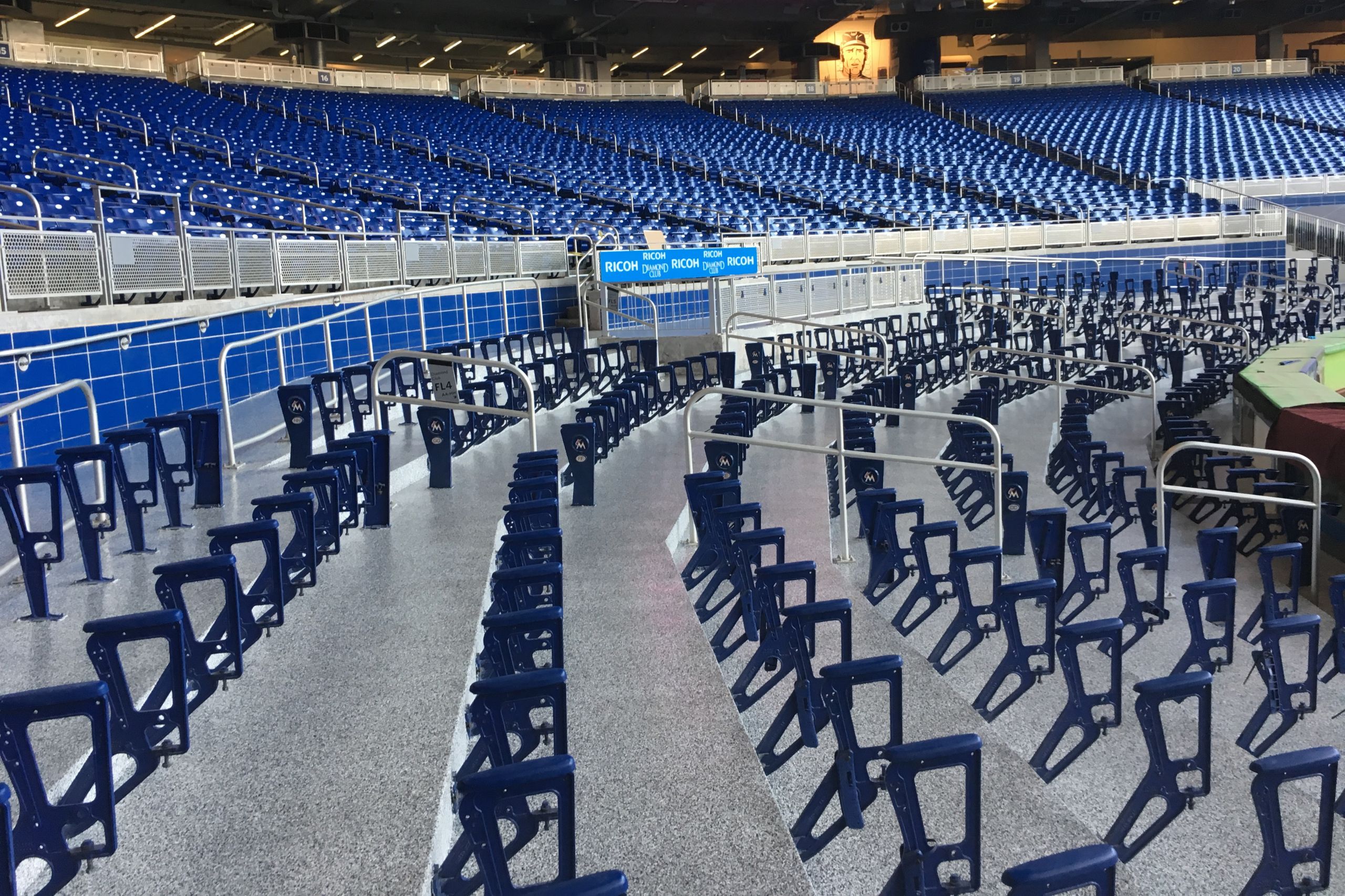 Rows of empty blue stadium seats, many with only their backs remaining, are set indoors on commercial and industrial flooring beneath artificial lighting.