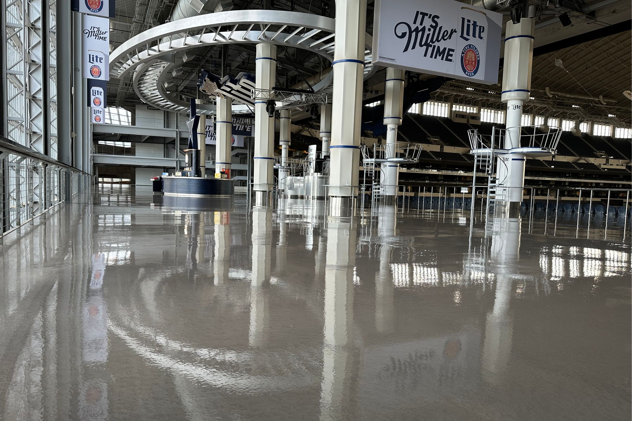 Interior of a large stadium concourse featuring commercial and industrial flooring, gleaming polished floors, support columns, and "It's Miller Lite Time" signs hanging from the ceiling.