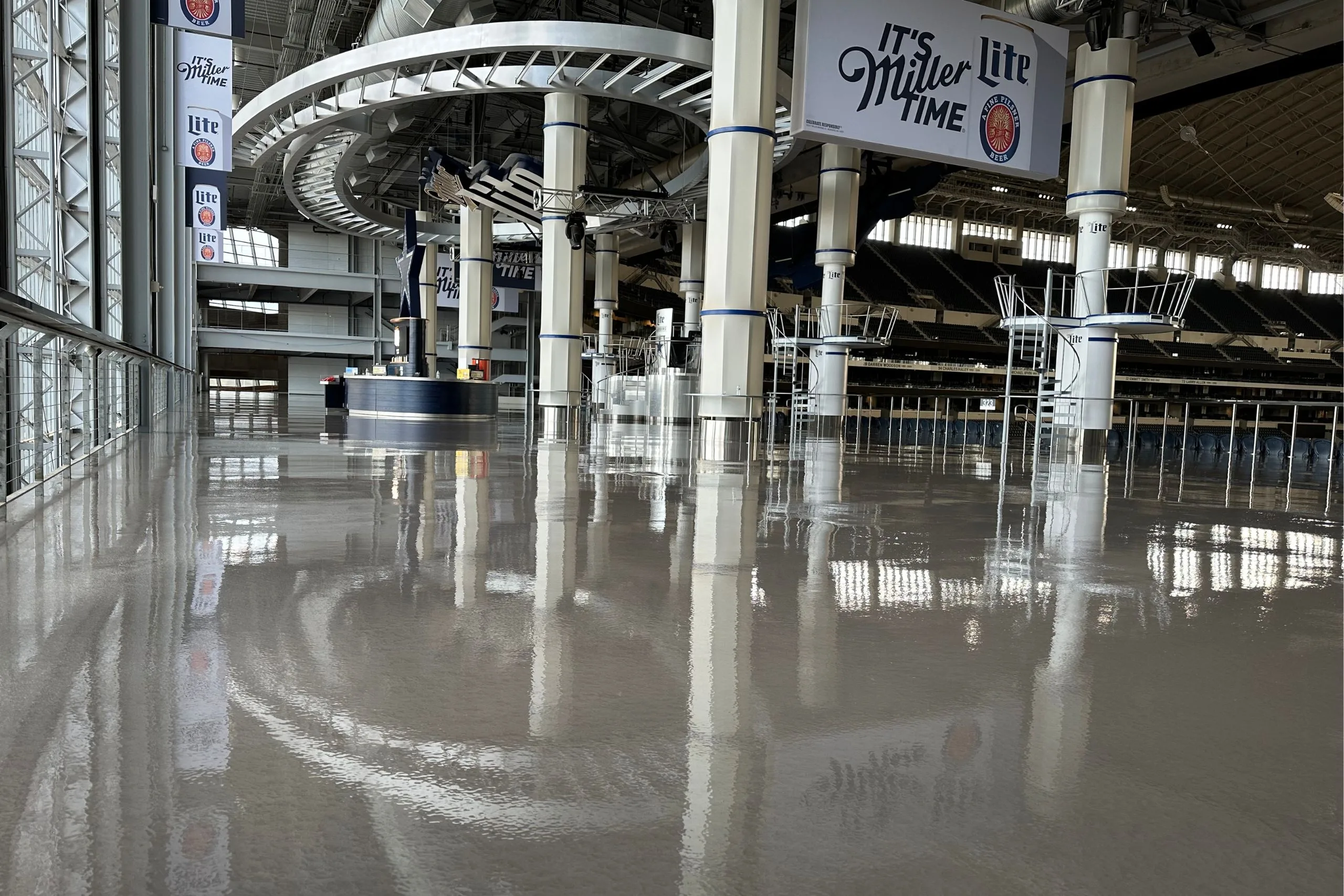 Interior of a large stadium concourse featuring commercial and industrial flooring, gleaming polished floors, support columns, and "It's Miller Lite Time" signs hanging from the ceiling.