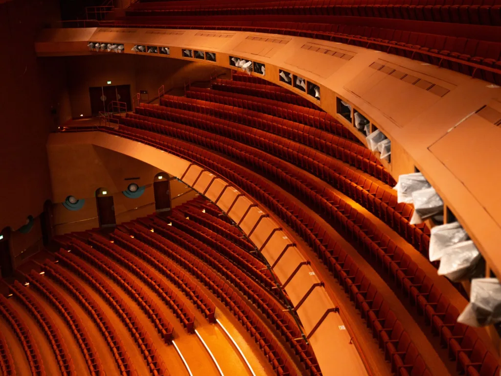 Rows of empty red theater seats curve around multiple levels in a large auditorium with soft lighting.