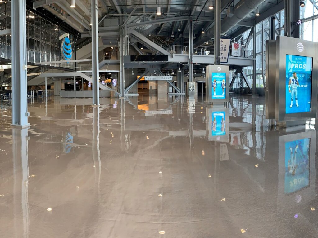 Large indoor stadium lobby with shiny polished floors, visible staircases, steel beams, and digital advertisement screens displaying sports content.