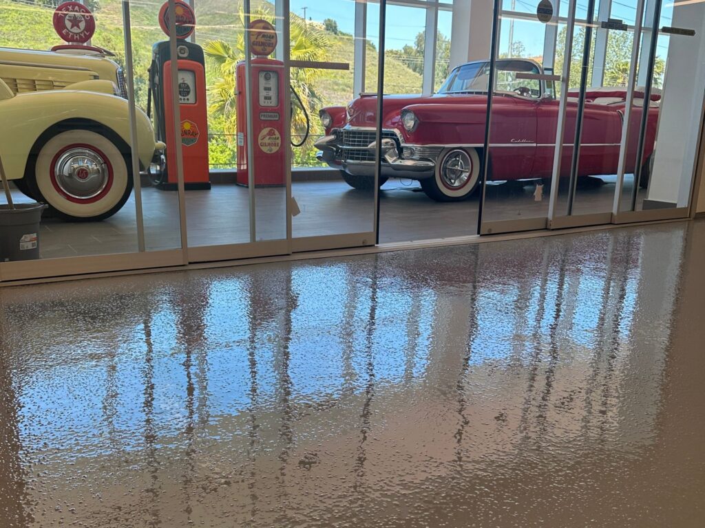 Two vintage cars and old-fashioned gas pumps are displayed behind glass walls, with their reflections visible on a shiny floor in the foreground.