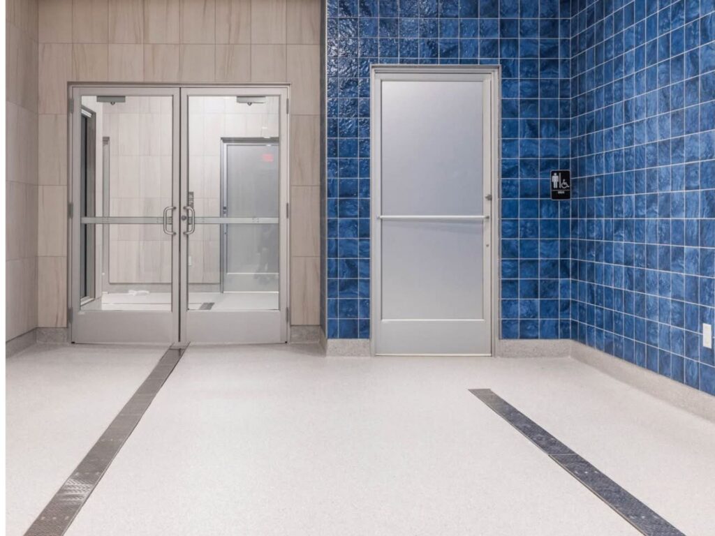 A hallway with white floors, blue tiled walls, a metal door, a glass double door, and a wall-mounted hand sanitizer dispenser.