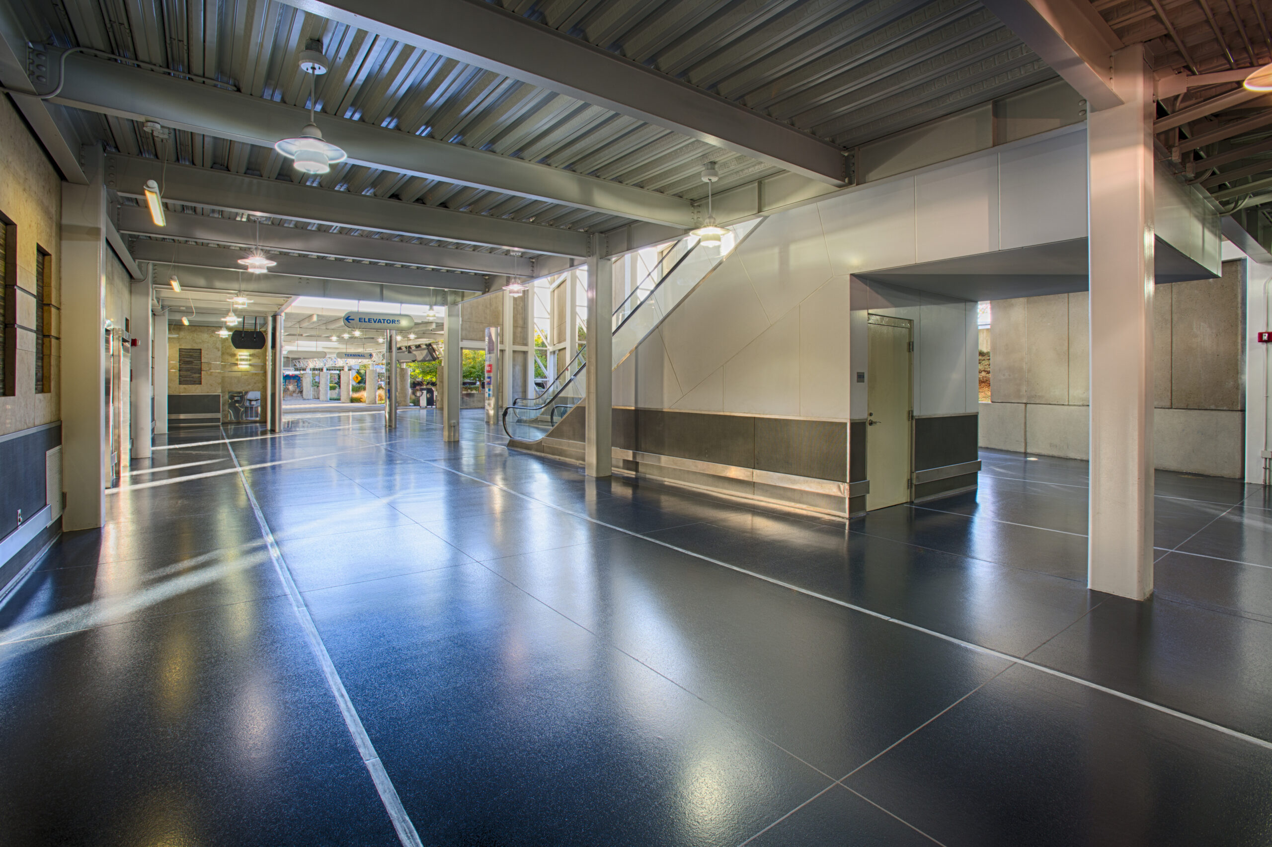 Modern building interior with shiny blue commercial and industrial flooring, exposed ceiling, large windows, glass doors, and an escalator in the background.