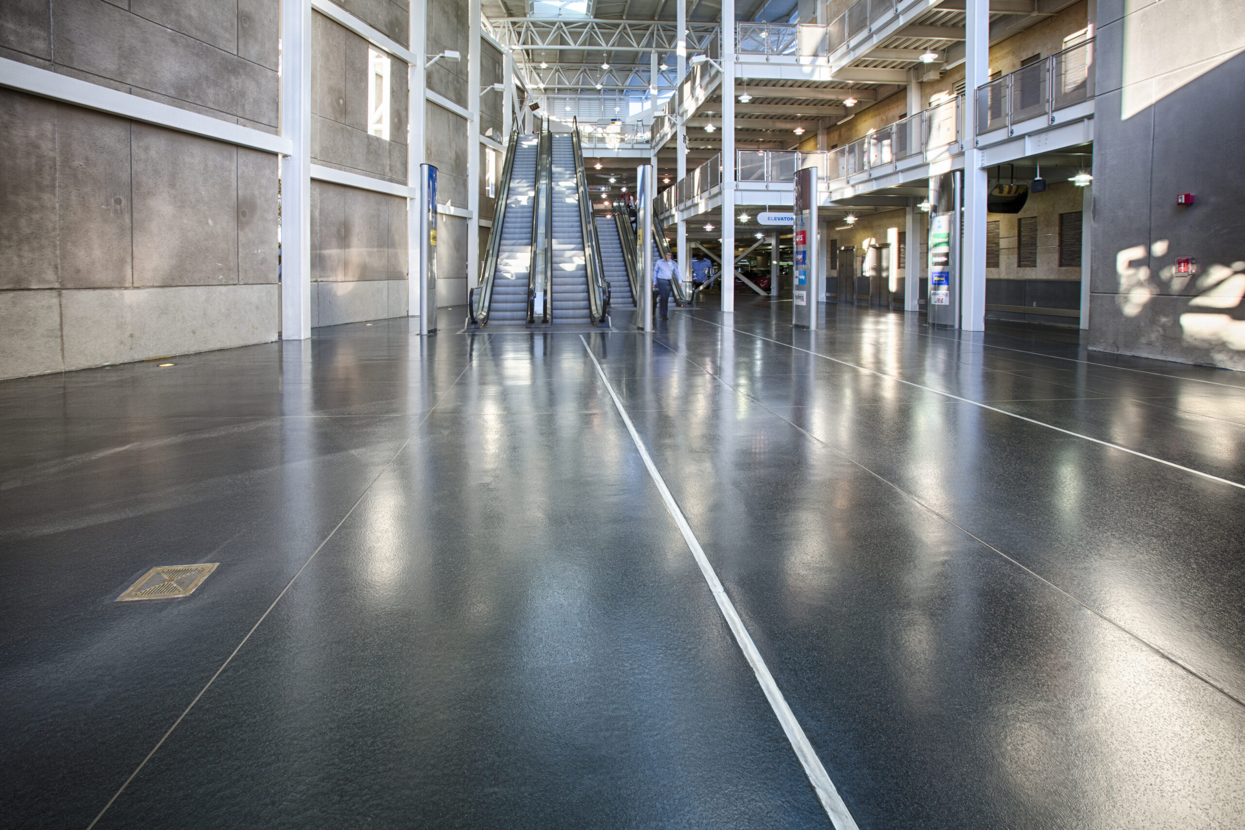 Spacious indoor atrium with polished commercial and industrial flooring, escalators, and a glass ceiling; natural light illuminates the modern architecture.
