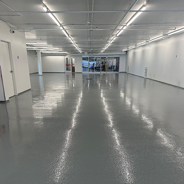Large empty room with glossy gray epoxy floor, white walls, and ceiling lights. Glass doors are visible at the far end of the space.