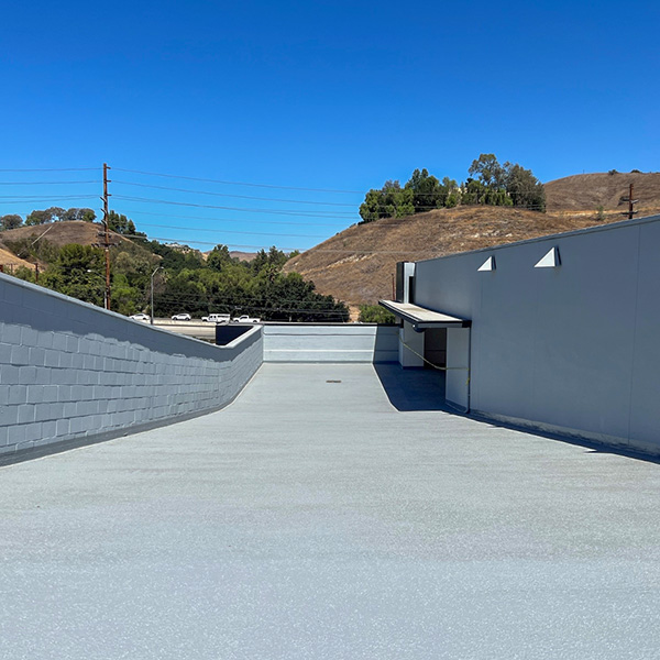 A flat, empty rooftop with smooth gray flooring next to a white building, overlooking hills and trees under a clear blue sky.
