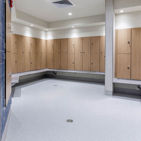 A clean, modern locker room with wooden lockers, gray benches, and overhead lighting. The floor is light-colored with a central drain.