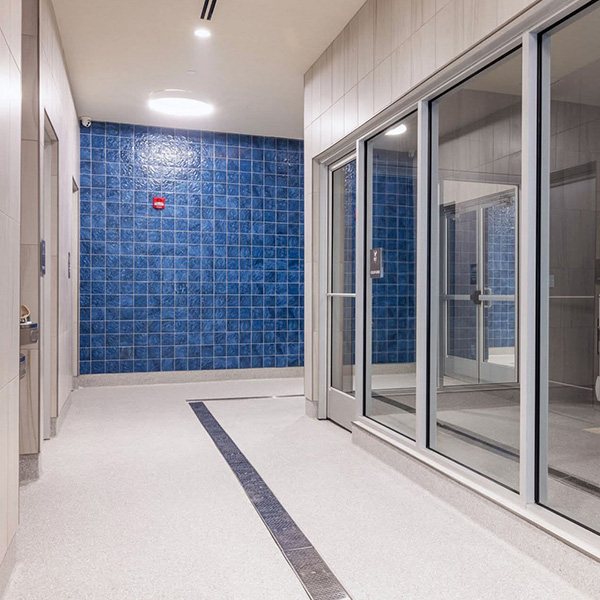 A modern hallway with blue tiled wall, gray flooring, and large glass windows on the right side. Ceiling lights illuminate the space.