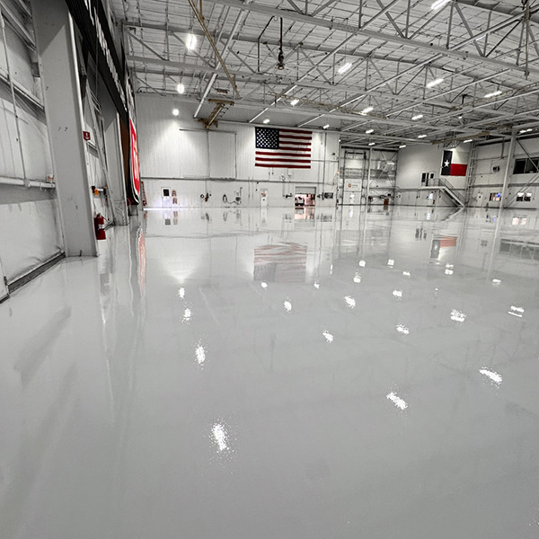Large, empty industrial warehouse with a shiny, polished floor, high ceiling, overhead lights, and American and Texas flags on the far wall.