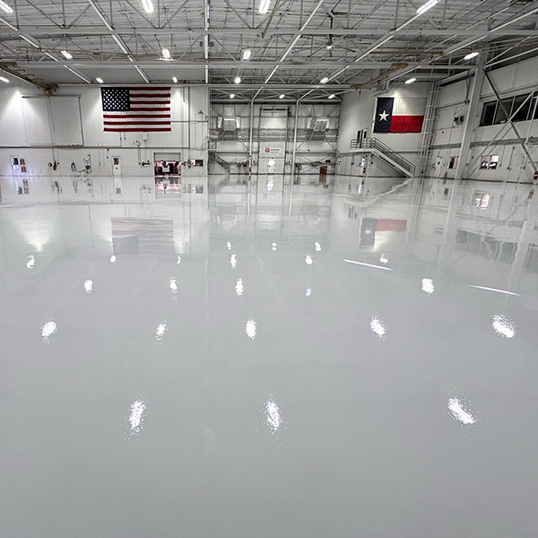 Large, empty indoor facility with polished floor, American and Texas flags on the walls, industrial lighting overhead, and visible doors and stairs along the sides.