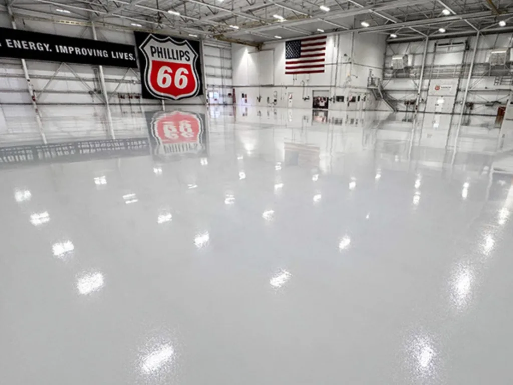 Large, clean, empty industrial hangar with polished white floors, an American flag, and a Phillips 66 sign on the wall. Bright overhead lights are reflected on the floor.