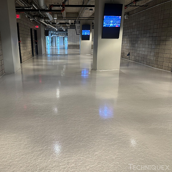 A brightly lit hallway with a glossy white floor, gray cinderblock walls, ceiling pipes, and screens mounted on pillars displaying a sports game.