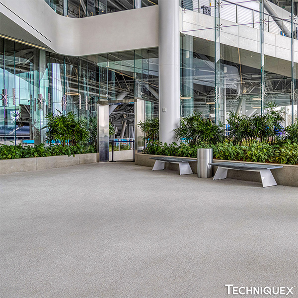 Modern indoor lobby area with large glass windows, greenery, two metal benches, a trash bin, and a visible upper floor walkway.