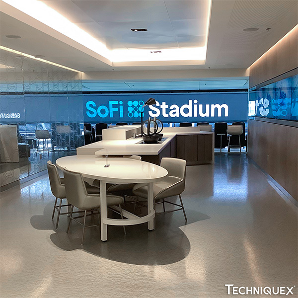 Modern lounge area with white tables and gray chairs, large SoFi Stadium sign on the glass wall in the background, and neutral-toned decor.