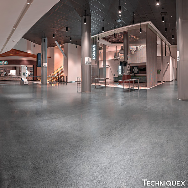 Spacious, modern indoor lobby with gray flooring, multiple pillars, ceiling lights, and illuminated signs; minimal furniture and a staircase visible in the background.