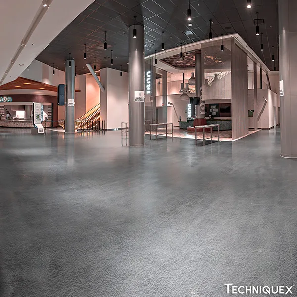 Spacious, modern indoor lobby with gray flooring, multiple pillars, ceiling lights, and illuminated signs; minimal furniture and a staircase visible in the background.