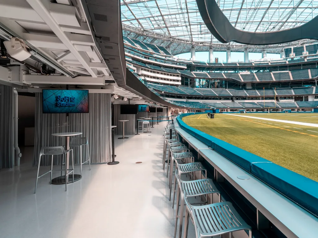 A modern stadium interior with tables, high chairs, and a view of the field; digital screens display "SoFi Stadium." The roof is glass, letting in natural light.
