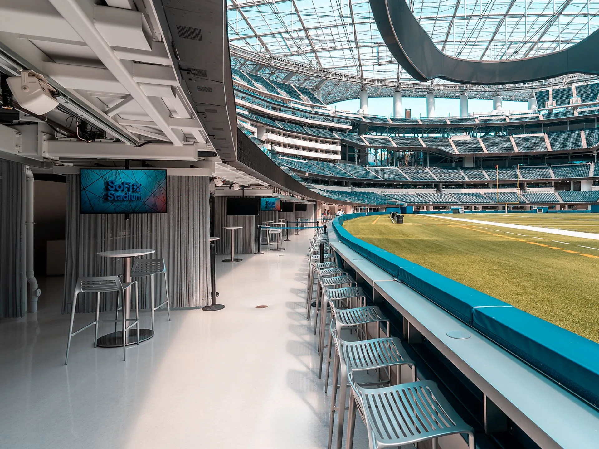 A modern stadium interior with tables, high chairs, and a view of the field; digital screens display "SoFi Stadium." The roof is glass, letting in natural light.