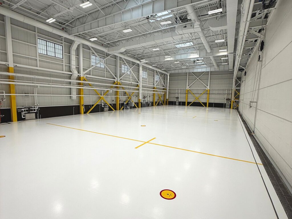 Large, empty indoor gymnasium with white floor, yellow lines, high ceiling, exposed beams, and structural supports. Bright lighting and visible HVAC ducts overhead.