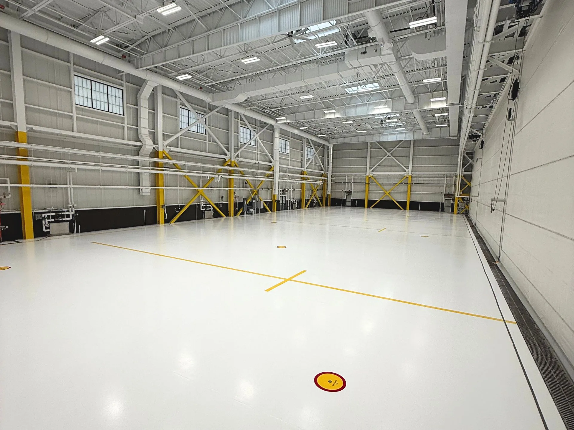 Large, empty indoor gymnasium with white floor, yellow lines, high ceiling, exposed beams, and structural supports. Bright lighting and visible HVAC ducts overhead.