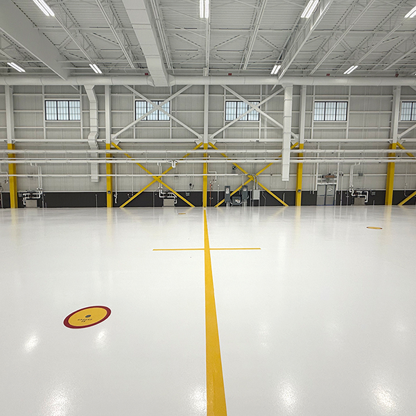 Spacious, empty industrial warehouse with white floors, yellow support beams, high ceilings, and large windows. A yellow line and a red circle are marked on the floor.