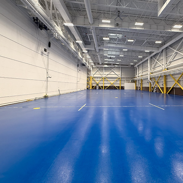 Large indoor gymnasium with a bright blue floor, white walls, high ceiling, and yellow support beams; no people or equipment visible.