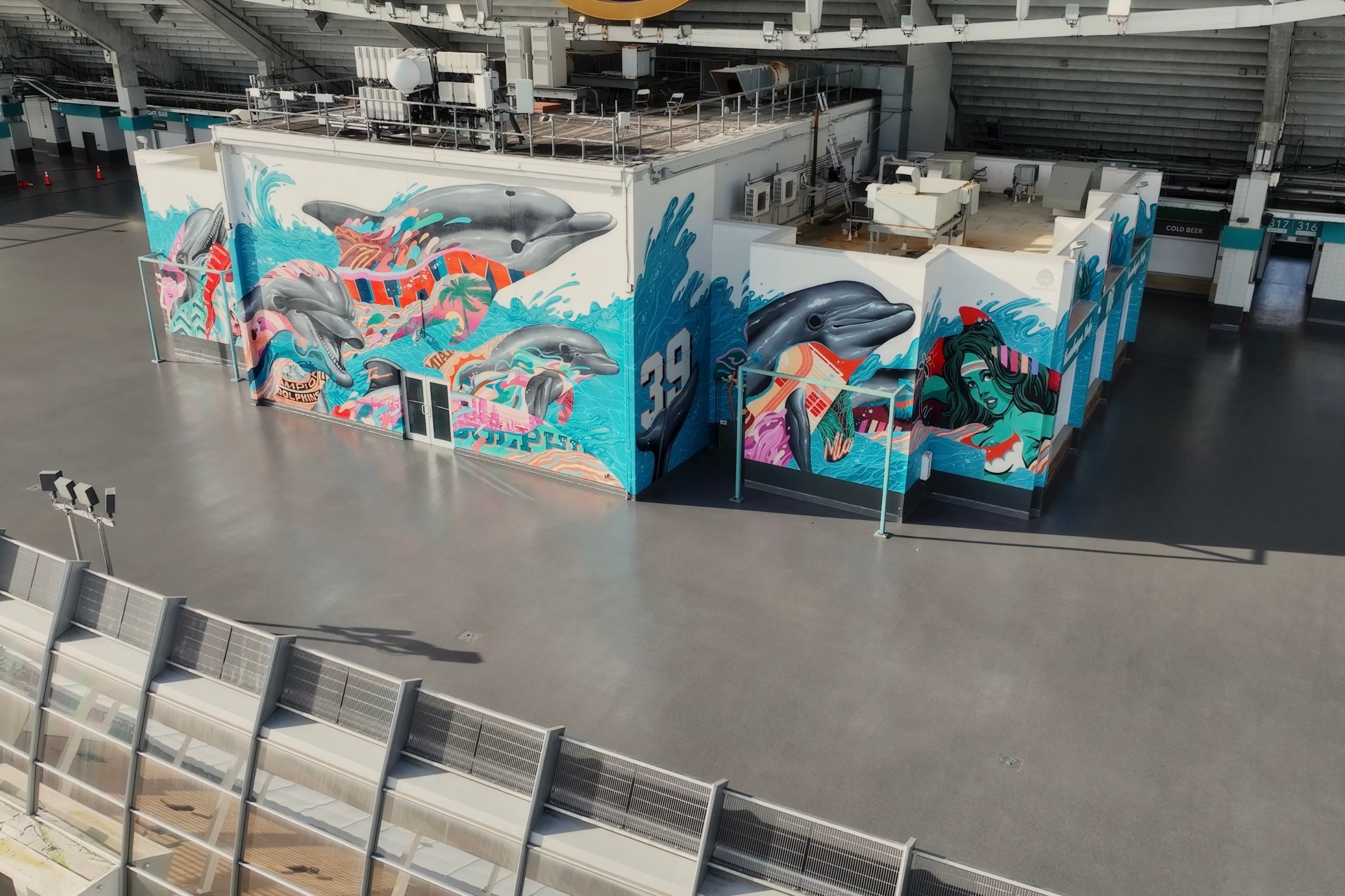 A building inside a stadium features commercial and industrial flooring, decorated with colorful murals of dolphins, ocean waves, and tropical patterns; the area around it is empty and spacious.