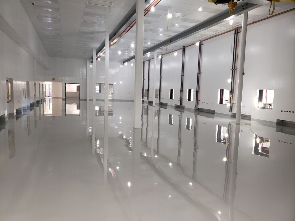 Large, empty industrial room with white walls, high ceiling, and glossy reflective floor, featuring multiple windows and vertical columns.