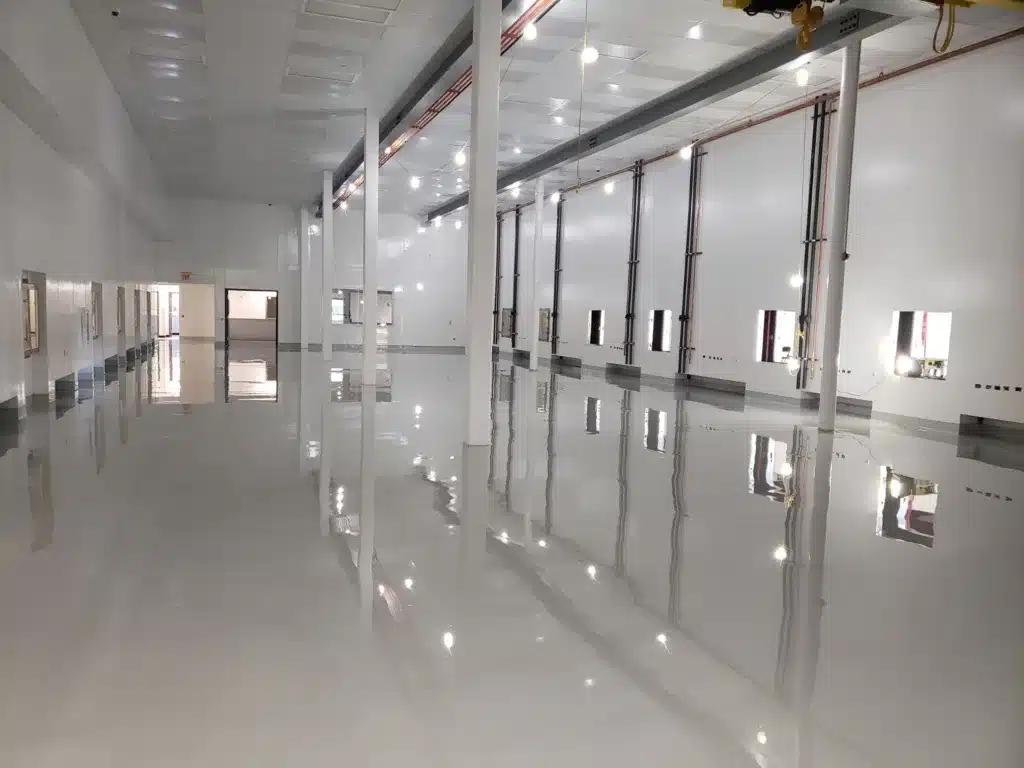 Large, empty industrial room with white walls, high ceiling, and glossy reflective floor, featuring multiple windows and vertical columns.