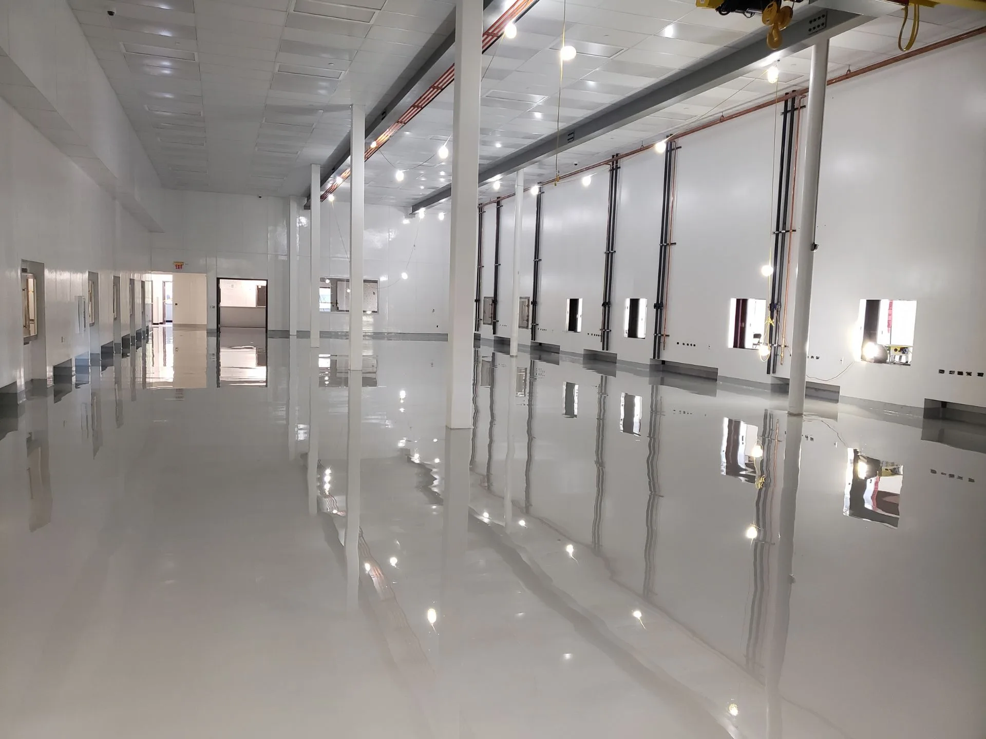 Large, empty industrial room with white walls, high ceiling, and glossy reflective floor, featuring multiple windows and vertical columns.