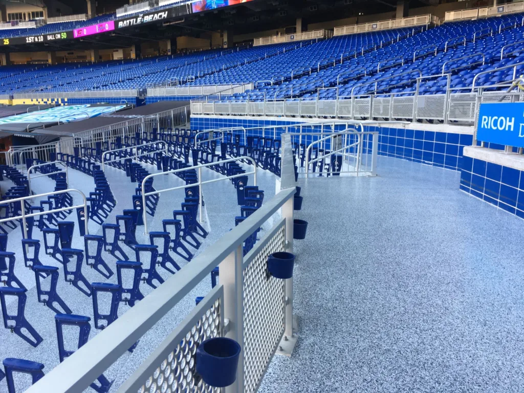 A stadium with empty blue seats and a wide, clean walkway bordered by metal railings. The scene appears brightly lit by daylight.