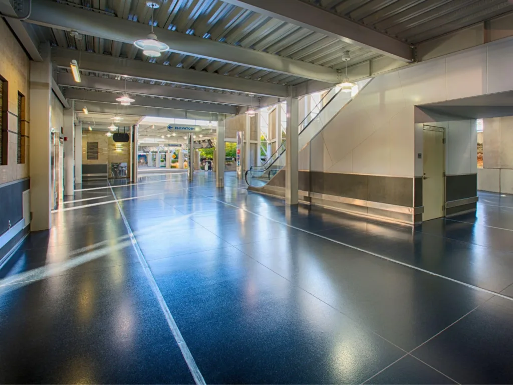 Spacious, modern indoor walkway with polished floors, an escalator, and glass doors leading to shops in the background. Ceiling lights and natural light illuminate the area.