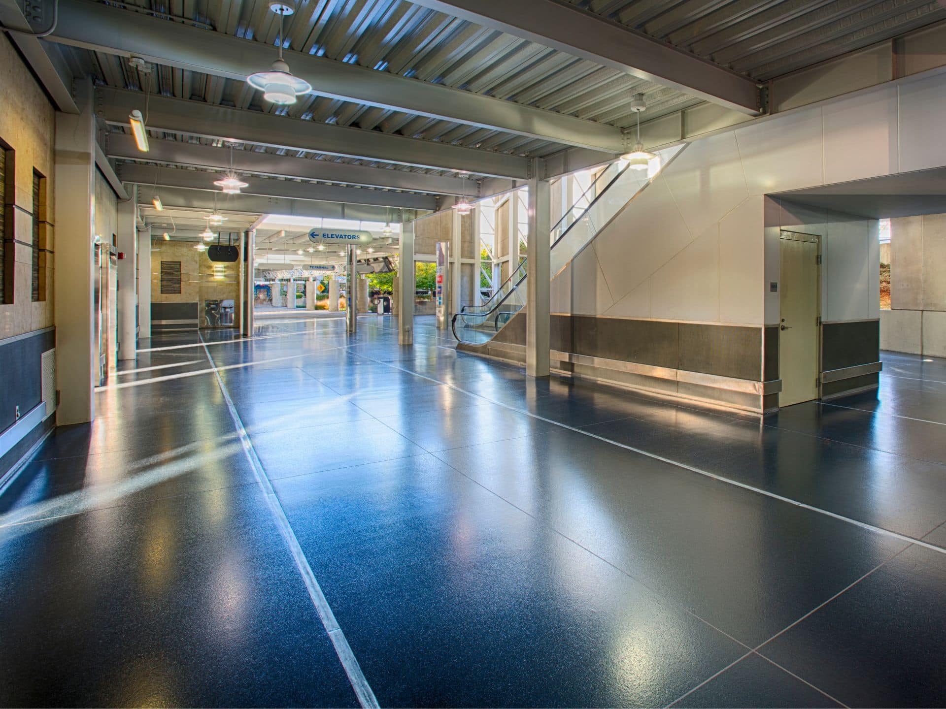 Spacious, modern indoor walkway with polished floors, an escalator, and glass doors leading to shops in the background. Ceiling lights and natural light illuminate the area.