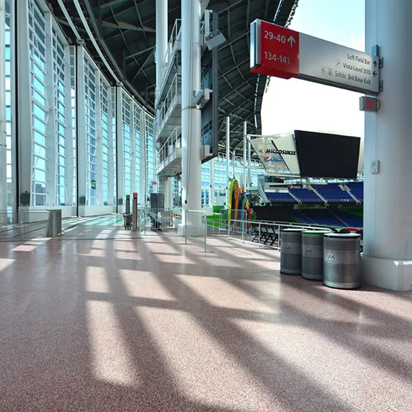 Interior view of a modern baseball stadium concourse with large windows, overhead signs, and three trash cans in the foreground.