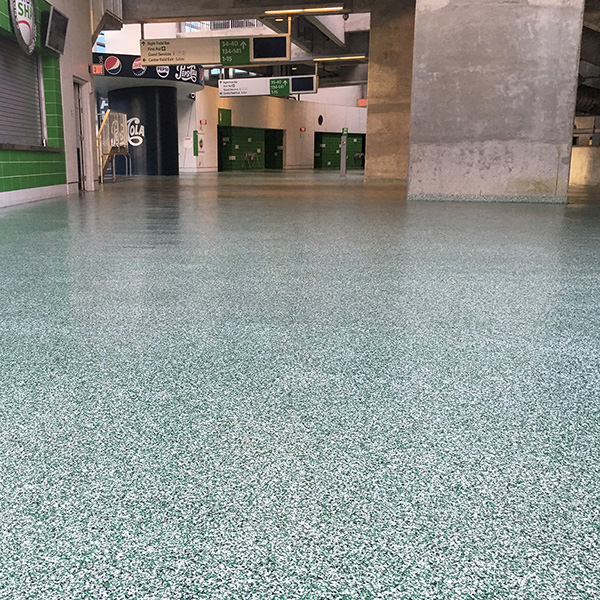 A wide view of an empty stadium concourse with green speckled flooring, concrete columns, closed concessions, and directional signs overhead.