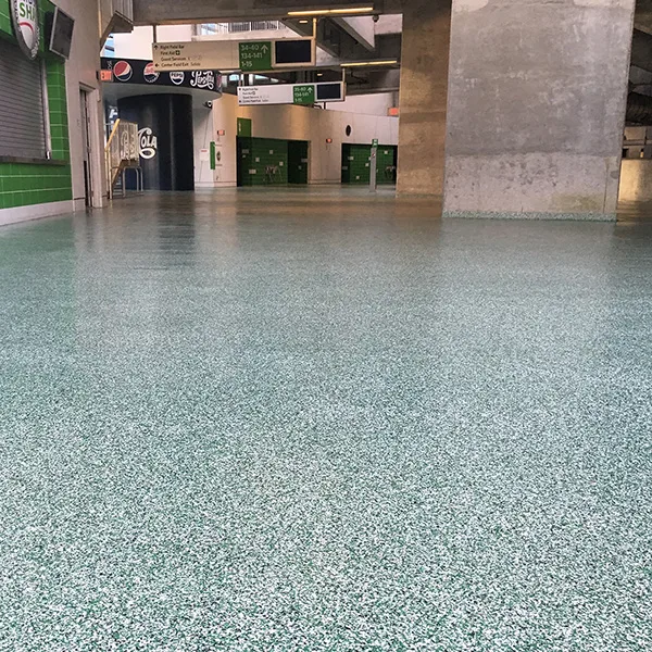 A wide view of an empty stadium concourse with green speckled flooring, concrete columns, closed concessions, and directional signs overhead.